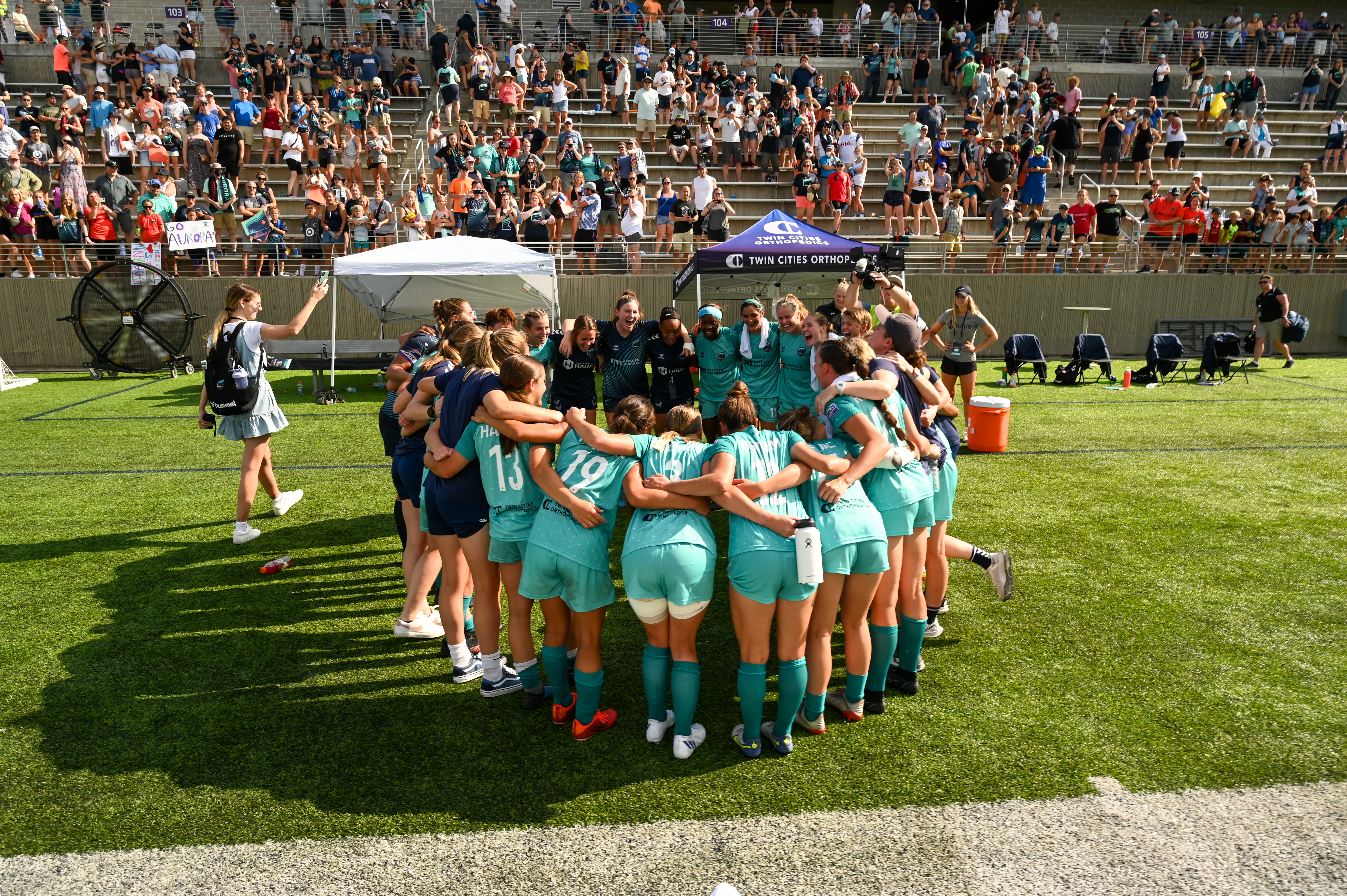 Players dressed in Minnesota Aurora FC’s turquoise jerseys in a huddle during a game in front of spectators in the stadium